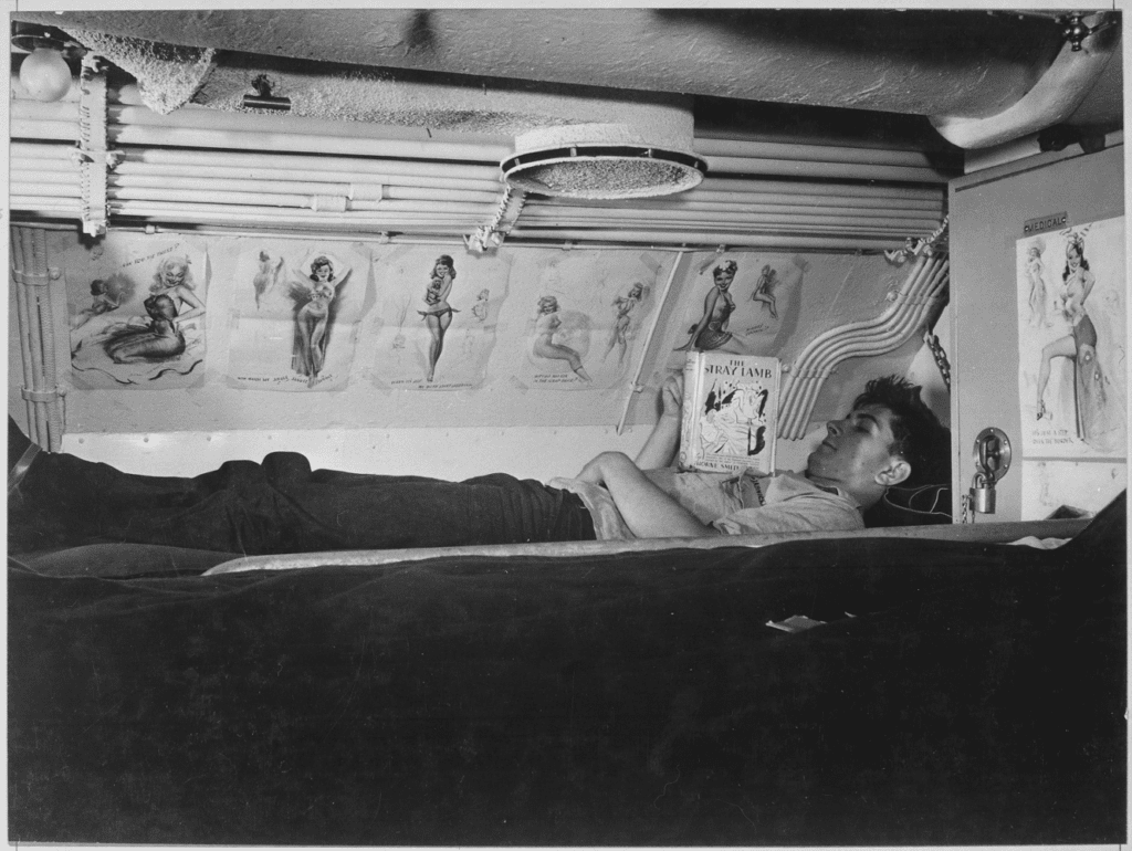 Sailor reading The Stray Lamb (1929) by Thorne Smith in his pinup-decorated bunk aboard USS Capelin (SS-289) at Naval Submarine Base New London in Connecticut, August 1943.