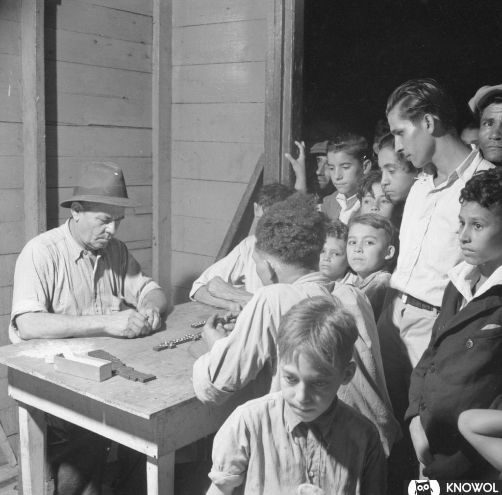 Two men playing a game of dominoes at a table while a group gathers around to watch.