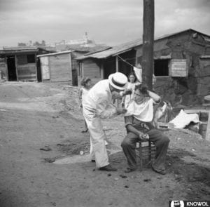 Barber cutting a man's hair outside, surrounded by shacks.