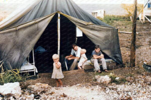 The photo shows a family outside a large military-style tent that appears to be their living quarters. In the foreground, a young child, likely a girl given the dress, stands on a rocky terrain, sucking her thumb, seemingly lost in thought. To the right, seated on the wooden steps of the tent, are three individuals who appear to be family members: a man with his arm around a small child and a boy sitting beside them, all looking weary or contemplative. The setting suggests a temporary, perhaps makeshift, living situation. The surrounding landscape is sparse and scrubby, indicating a rural or undeveloped area.